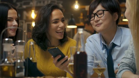 Young Asian Man Shows Interesting Stuff on His Smartphone to His Friends while They Have Good Time in Bar. They Laugh, Joke, Drink in Stylish Hipster Bar Establishment. 