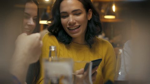 Beautiful Hispanic Woman Uses Smartphone While Talking and Having Fun with Her Friends in the Bar. They Laugh, Joke, Drink in Stylish Hipster Bar Establishment. : film stockowy