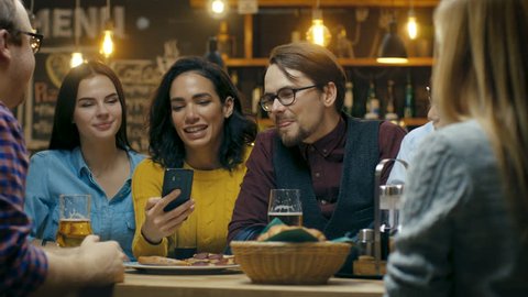 Beautiful Hispanic Woman Shows Interesting Stuff on Her Smartphone to Her Friends while They Have Good Time in Bar. They Laugh, Joke, Drink in Stylish Hipster Bar Establishment. 