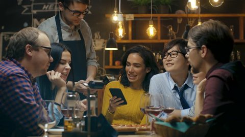 In the Bar Waiter Holds Credit Card Payment Machine and Beautiful Woman Pays for Her Order with Contactless Mobile Phone Payments System. She's Surrounded by Dear Friends and Has Time of Her Life. Sho