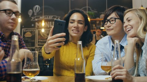 In the Bar/ Restaurant Hispanic Woman Makes Video Call with Her Friends. Group Beautiful Young People in Stylish Establishment. 