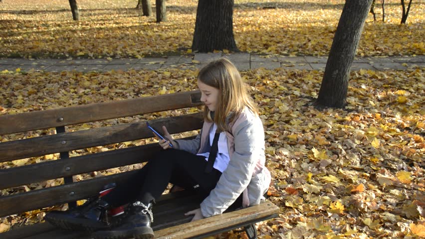 beautiful girl on a bench in an autumn park, holding a smartphone in her hands. girl smiling, waving her hand and laughing. the child enjoys good weather and communication with friends and family
