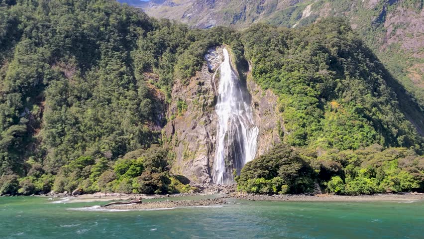 Bowen Falls (Hineteawa in Māori language) with Mitre Peak in Milford Sound (Piopiotahi) with surrounding mountains. A sunny blue sky day in Fiordland National Park on the South Island of New Zealand Royalty-Free Stock Footage #3416186015