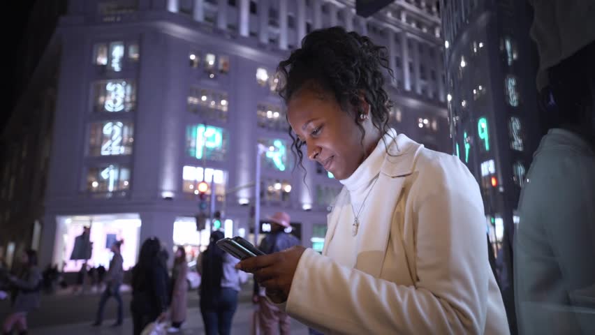 Young latina woman waits sitting for her date in night illuminated winter city center. Smiling afro female types on mobile phone in dating app. Generation z black people and social relationships. Royalty-Free Stock Footage #3416201153