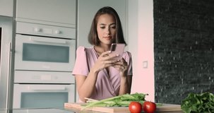 Young attractive female holding modern smart phone, distracted from healthy vegetable salad preparation. Food blogging, on-line cookbook recipes application usage, chatting while cooking in kitchen