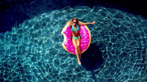 Young happy woman relaxing on inflatable pool toy in blue swimming pool on sunny day waiving at camera