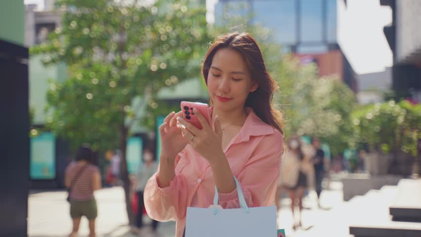 Asian beautiful women looking on map while walk in department store. Attractive young female shoper holding shopping bag and using smartphone with happiness enjoy purchasing in shopping mall center.