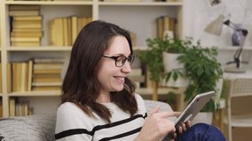Portrait of a young woman with glasses sitting in an armchair, smiling and using a tablet indoors. Wireless Internet and mobile technologies - Powered by Shutterstock - Get 15% off with code: PIKWIZARD15
