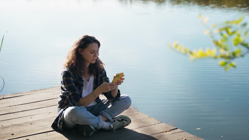 A young Caucasian woman uses her phone while sitting on a wooden pier on a lake on a sunny day.