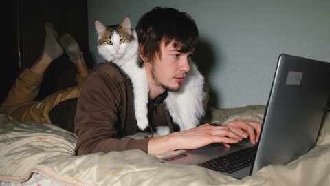 A young man working at the laptop on the bed with his cat, closeup.