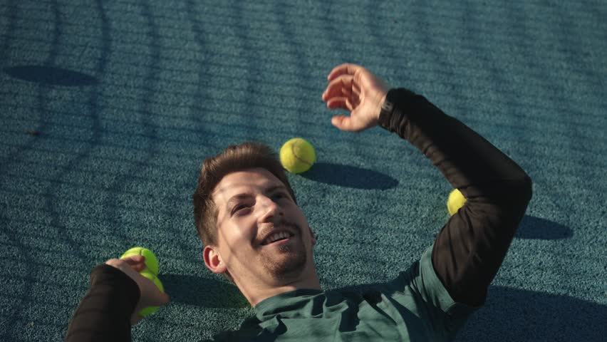 A bearded model with fair skin lies on the blue tennis court, tossing a tennis ball into the air with a smile, while a shadow falls across his face.
