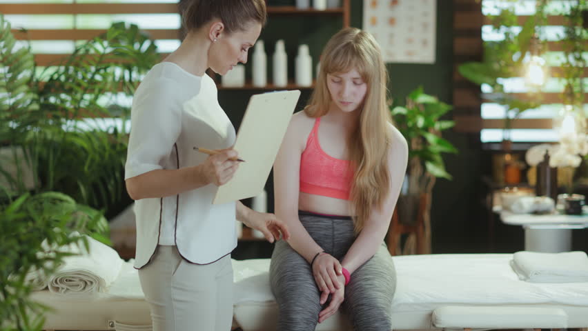 Healthcare time. concerned massage therapist woman in massage cabinet with clipboard and teenage client listening to a clients complaint of leg pain. Royalty-Free Stock Footage #3417339943