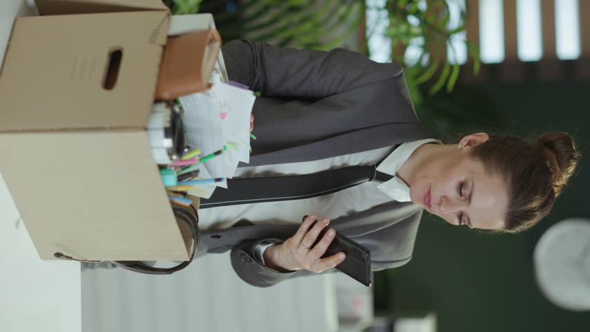 New job. stressed modern woman worker in modern green office in grey business suit with personal belongings in cardboard box speaking on a smartphone. Royalty-Free Stock Footage #3417490013