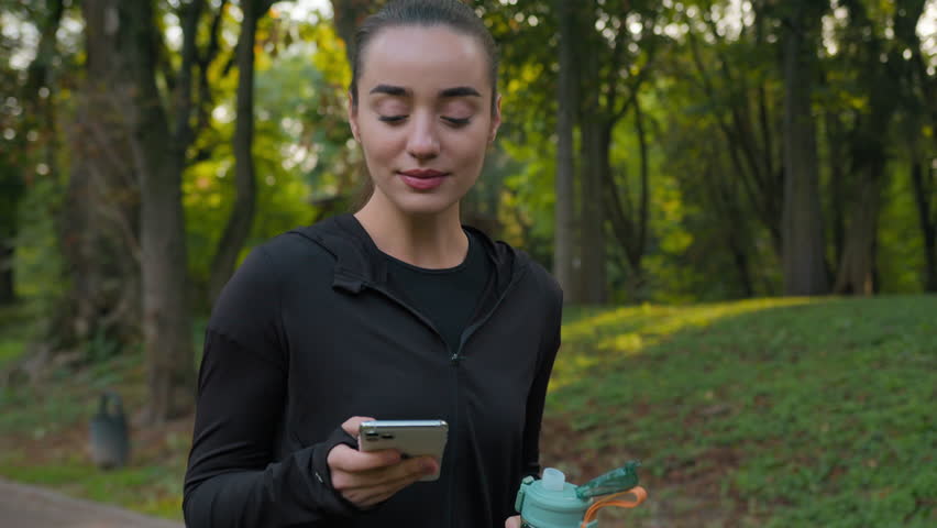 Fitness woman Caucasian sporty girl walking in city park with sport bottle drinking water using smartphone check mobile phone social media happy female young blogger training healthy workout outdoors