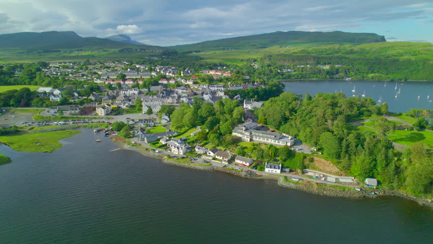 AERIAL: Peaceful bay by the picturesque island town of Portree on a sunny day. Charming port city in sheltered coast of a hilly and green Isle of Skye. Magnificent landscape in northern Scotland.