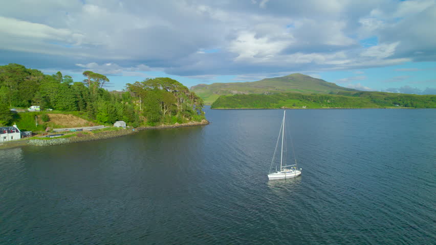 AERIAL: White sailboat dropped anchor in a peaceful bay on beautiful Isle of Skye near the capital island town of Portree. Magnificent coastline of a hilly and green island in the north of Scotland.