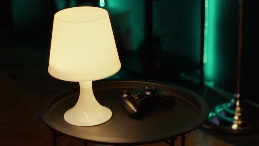 Panning shot of gaming console controller sitting on nightstand in neon illuminated living room. Close up of game system gamepad joystick on small table next to lit lamp in dark apartment