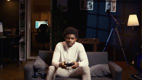 African american man playing shooter videogame in cozy living room, holding controller. Gamer participating in PvP online multiplayer game using console system, relaxing at home
