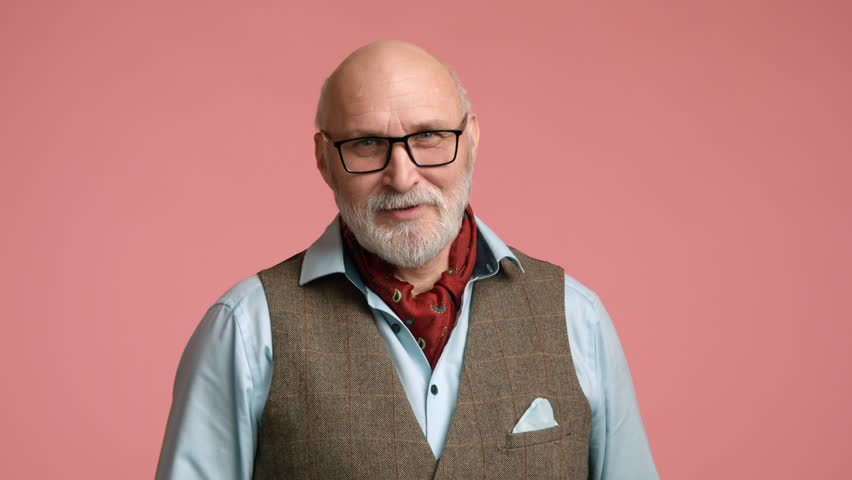 A senior man in a blue shirt and tweed vest is greeting with a raised hand, showcasing a friendly demeanor on a pink background. Camera 8K RAW. 