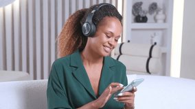 Young positive energetic African American woman meloman with headphones on ears listens to music and holds phone to select tracks in apps for online streaming of clips sits on sofa inside apartment.