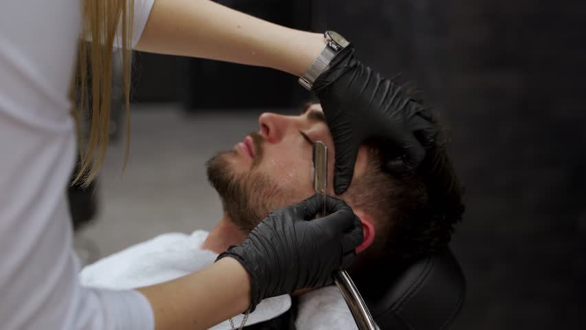 Professional hairstylist shaves client with straight razor in upscale barbershop. Man relaxes during facial grooming session. Skilled female barber gives beard trim, luxury grooming experience.