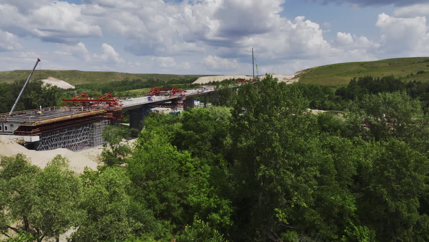 Aerial view of bridge construction from side of dense forest in sunny day. Narrow river that flows in middle of wooded area under bridge Royalty-Free Stock Footage #3417787823