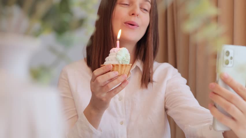 Cheerful brown haired woman with cake celebrating birthday on video call singing happy birthday song congratulating her friend with her holiday enjoying festive mood