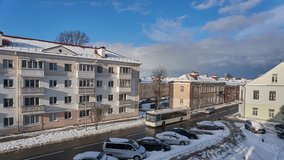 A quiet and peaceful street in the city in winter. Timelapse video clouds float over a small multi-storey building and the roadway. The road and cars are covered with snow.