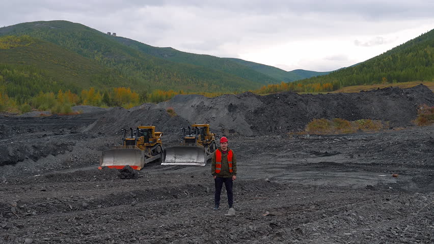 Miner standing in front of the industrial excavation equipment at the quarry. Miner worker observing the industrial equipment gathering rocks. Industrial mining equipment operator wearing a hard hat.