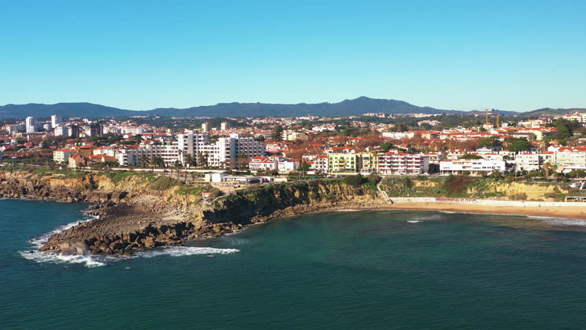 Aerial drone shot over turquoise sea and waves on the Atlantic ocean under blue sky, overlooking the cityscape, a beach and coast of Sao Pedro do Estoril on sunny day in Cascais, Portugal