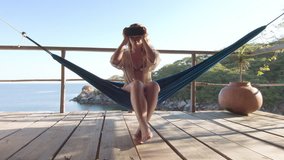 4k Imagination concept: Adult travel woman at exotic resort sitting on hammock, wearing virtual reality headset, watching 360 video experience at beach hut bungalow on holiday.