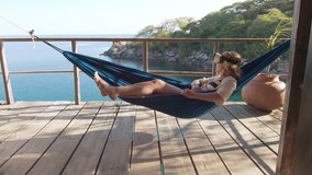 4k Imagination concept: Adult travel woman at exotic resort relaxing on hammock wearing virtual reality headset, watching 360 video experience at beach hut bungalow on holiday.