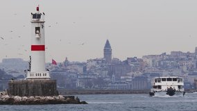 This video captures a vibrant coastal scene dominated by the Galata Tower rising from the misty city backdrop, juxtaposed with a foreground of a stark white lighthouse with red accents. A ferry, a
