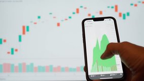 A young man hand scrolling on smartphone screen stock market  trading charts analyst.  On background a big screen with financial graphics - Powered by Shutterstock - Get 15% off with code: PIKWIZARD15
