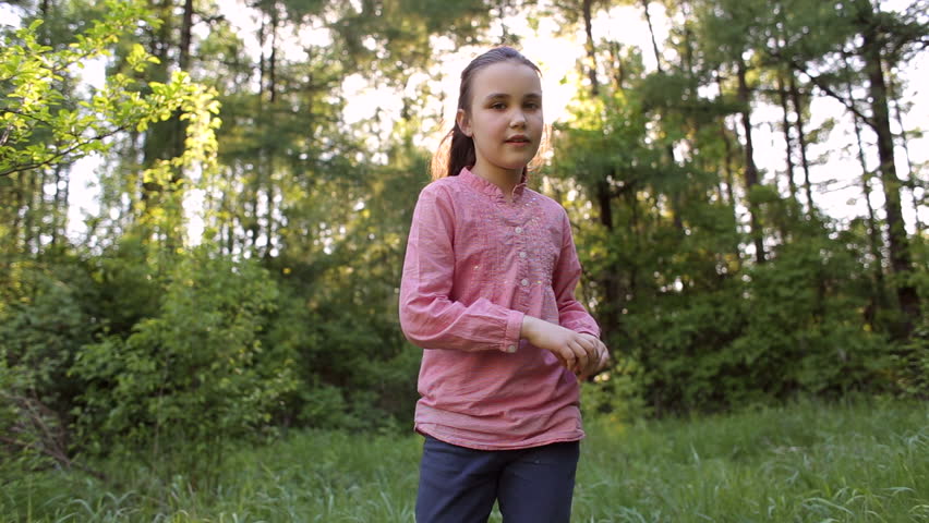A cute happy teenage girl in a pink shirt is spinning around herself in the park amid the setting sun.
