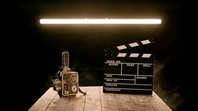 A clapper board and vintage video camera on a wooden table enveloped in smoke. Cinematography equipment in a studio on a black background illuminated by a neon lamp.
