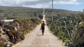 Popeye Village Exploration: 4K Shot of a Young Woman on a Dirt Road with Cactuses in Malta; Unveiling Charming Architecture, Rich History, and Seaside Beauty Near Iconic Film Set. - Powered by Shutterstock - Get 15% off with code: PIKWIZARD15