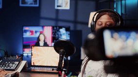Woman hosting podcast, saluting audience while recording entertaining discussion in apartment studio. Happy presenter uses professional camera to produce comedy internet show, greeting viewers