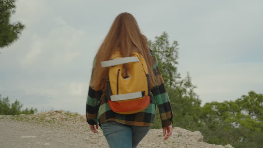 Ascent to the mountain, a woman with a bright backpack approaches the edge of the cliff and admires the treetops and the sea in the distance.
