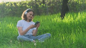 Happy carefree woman wearing headphones listens to music podcast sitting on green grass outdoors. Young beautiful girl enjoying good sunny day in the country. Handheld video 4K.