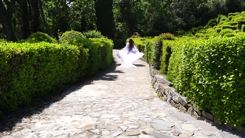 Happy Woman run in the park among lush green bushes, slow-motion. Girl in white flying dress with long Blond hair in the garden. Slim lady
