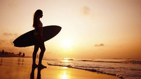 Tourist woman surfer in swimsuit with surfboard stands on ocean sandy beach at sunset admires waves. Summer sport activity on tropics resort. Sporty girl. Extreme water sport surfing on vacation. - Powered by Shutterstock - Get 15% off with code: PIKWIZARD15
