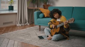 Young girl learning to play guitar. African american teenager girl study to playing the guitar see in laptop sitting on floor at home. Online-education, musician art, hobby practice concept.