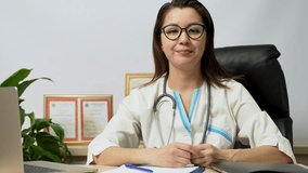Smiling young woman doctor cardiologist wearing white medical coat and stethoscope showing hands heart shape looking at camera at hospital office, close-up 4k slow motion