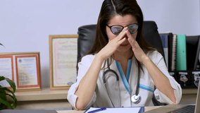 Stressed frustrated woman doctor wearing white coat with stethoscope covering face with hands, sitting at work desk in hospital.