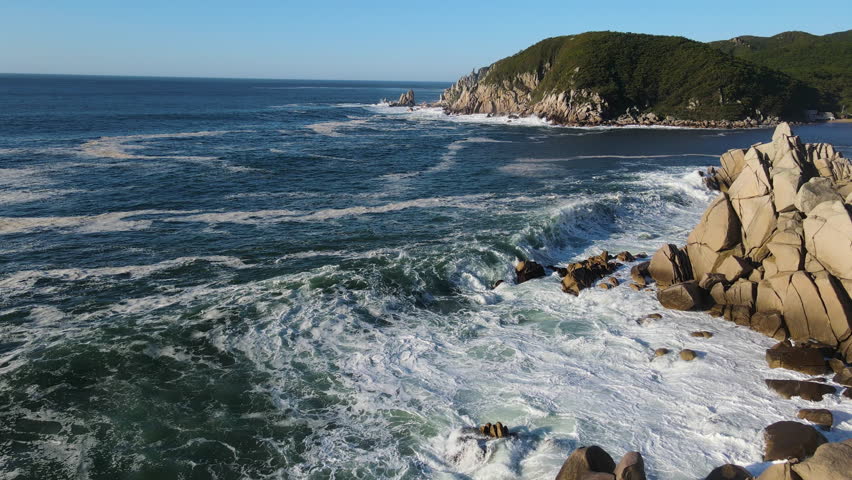 Drone slow motion of huge waves crashing on the seashore Sunny summer morning Rocky shore with rocks covered with green forest. High quality 4k footage