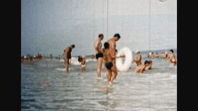 People crowd relax on sea beach in sunny summer. Parents, children with inflatable rings enjoy vacation in ocean shore. Many people in water. City public beach. 1980s archive. Old film. Archival video
