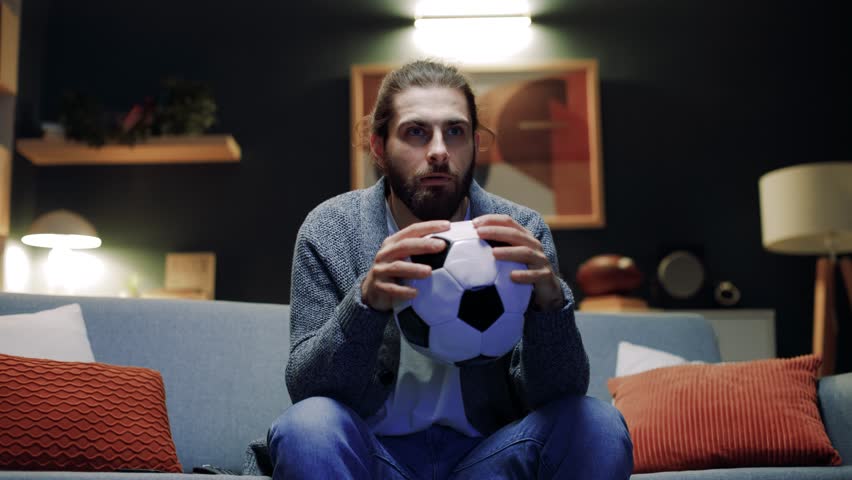 Male observing football game. Concentrated man holding pillow in shape of soccer ball. Male watching football match. Man cheering up for favorite team. Male nervous for match final.