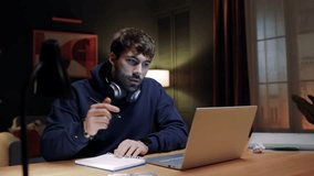 Man sitting at table with headphones on neck. Bearded male looking at laptop screen. Man holding pen and making notes. Male writing in notebook. Handsome man looking for inspiration.