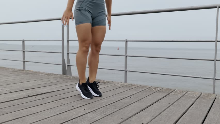 Determined and motivated young woman, 25, exercises outdoors on a cloudy day, performing Floor Touch Squat Jacks Royalty-Free Stock Footage #3418947711
